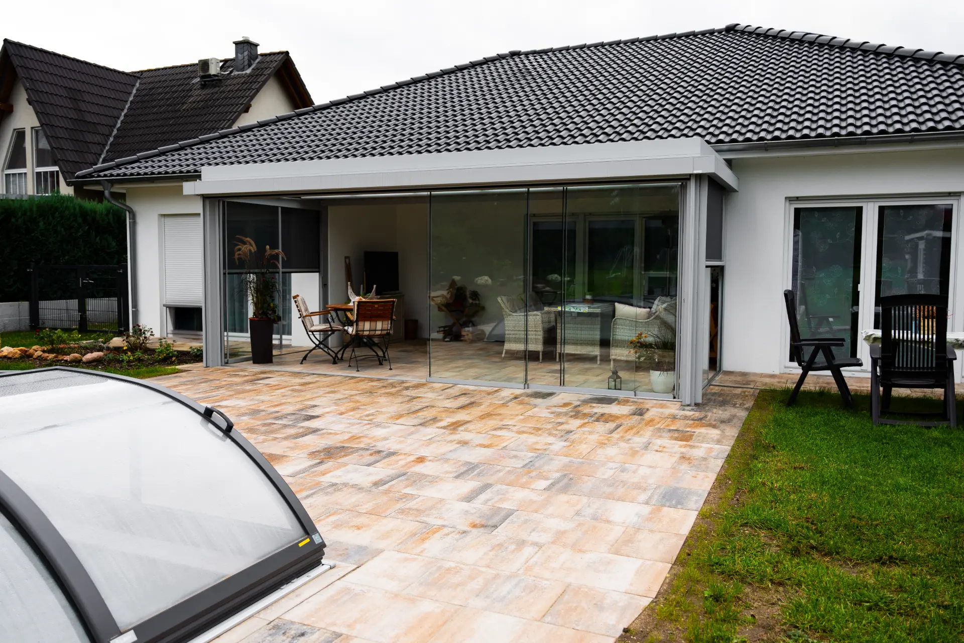 Terrassendach mit Glasschiebewand an einem weißen Bungalow, davor eine gepflasterte Terrasse