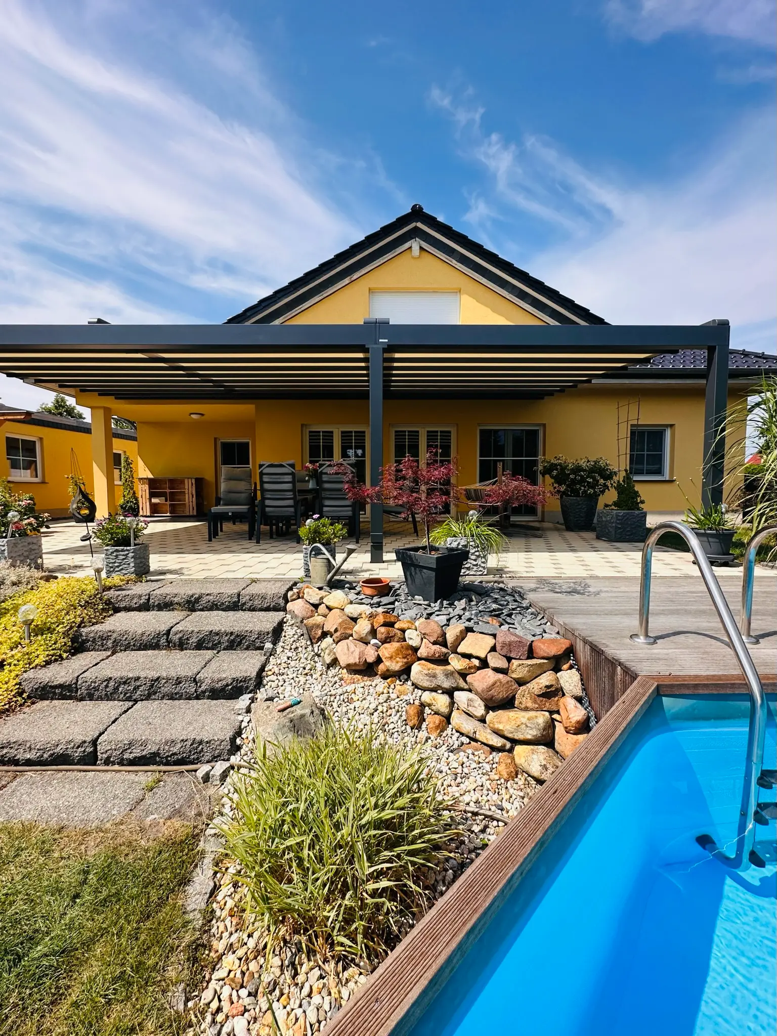 Lamellendach an einem gelben Bungalow, davor Pool mit Steingarten